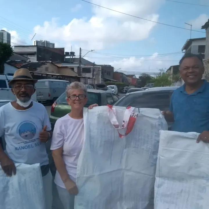 Três pessoas segurando grandes sacos brancos de material reciclável, em frente a carros, numa rua.