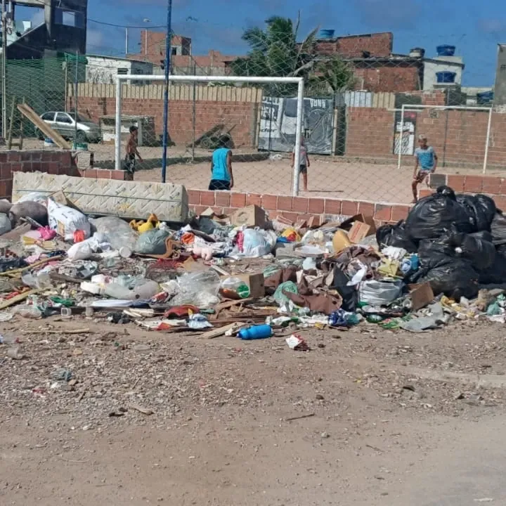 Entorno de uma comunidade: campo de futebol e pessoas ao fundo, com acúmulo de resíduos na frente.